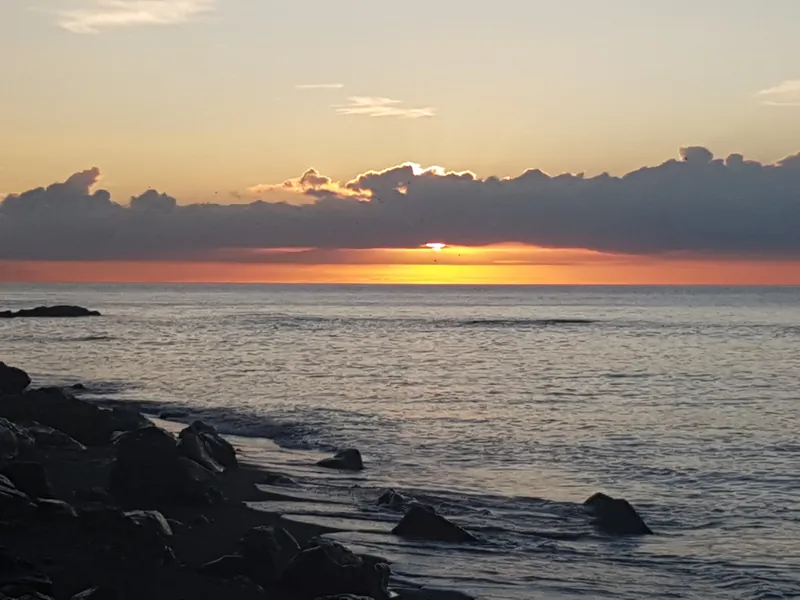 Amanecer con el sol asomando bajo una franja de nubes sobre el mar y rocas en la orilla