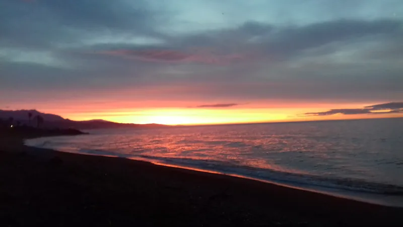 Cielo rojizo y naranja al amanecer sobre una playa de arena oscura con montañas al fondo