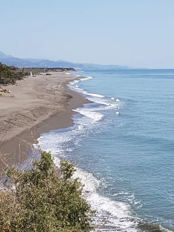Playa de arena y mar azul vistos desde lo alto de un acantilado, con montañas lejanas
