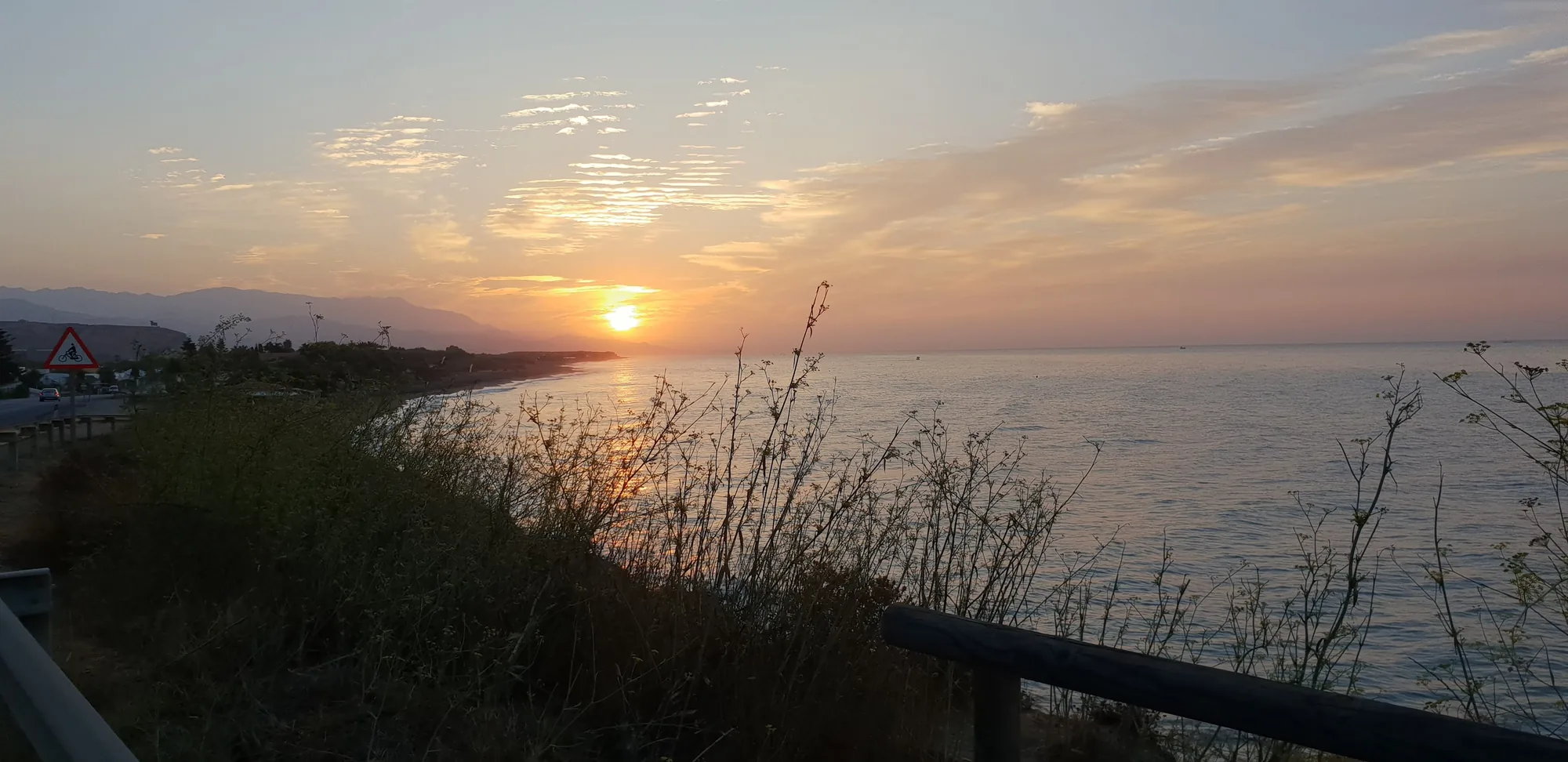 Sol bajo sobre el mar en una costa con vegetación y una señal de carretera en Valle-Niza