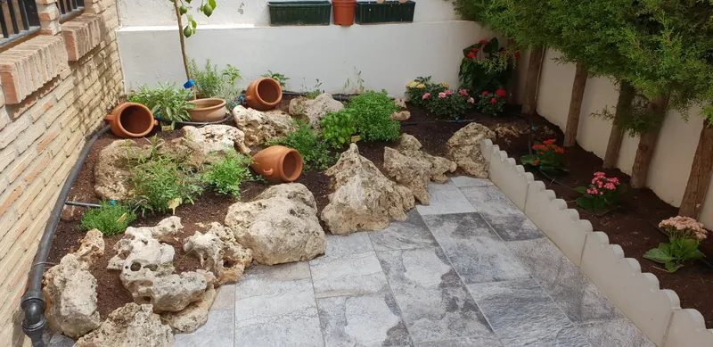 Rincón de jardín con rocas, macetas de barro y plantas junto a un muro blanco