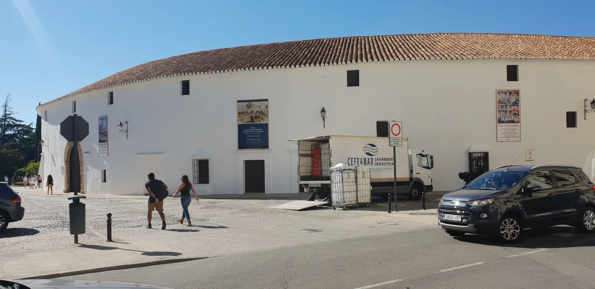 Plaza con un edificio blanco de tejado de teja, gente paseando y un coche aparcado en Ronda