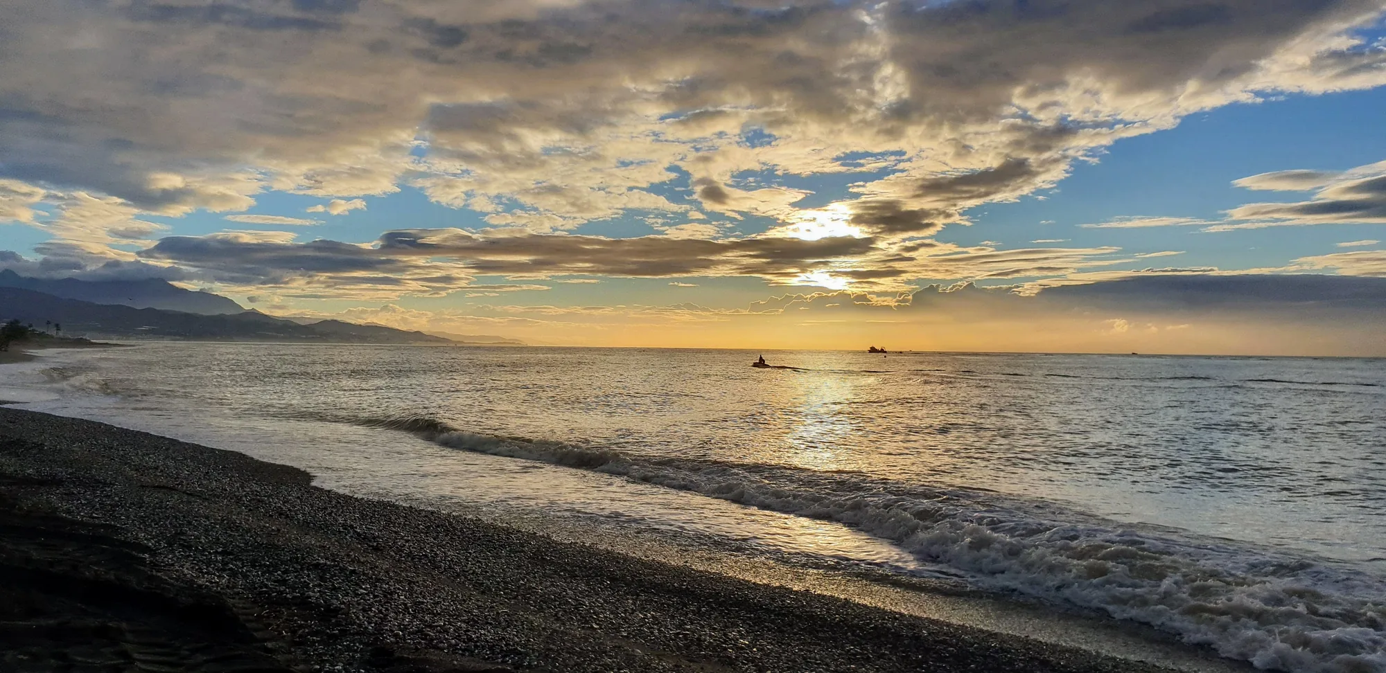 Olas llegando a una playa oscura bajo un cielo de nubes con el sol entre ellas en Valle-Niza