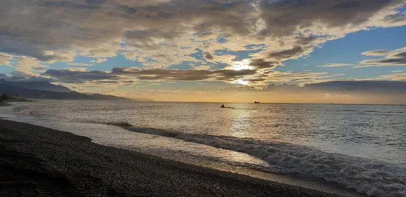 Olas llegando a una playa de cantos bajo un cielo con el sol entre nubes