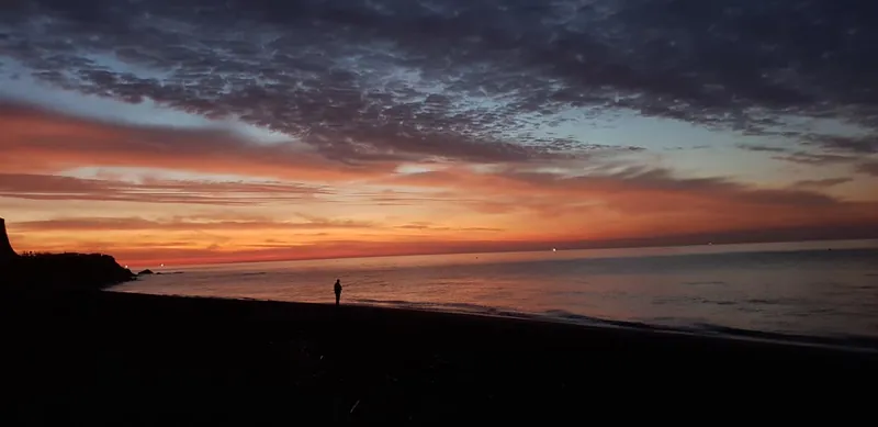 Una persona pequeña en la orilla bajo un cielo de nubes rojizas al amanecer