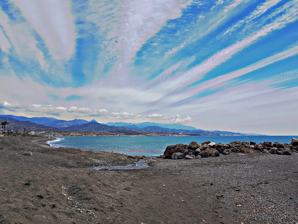 Cielo azul surcado por nubes alargadas sobre una playa con rocas y montañas en Torre del Mar