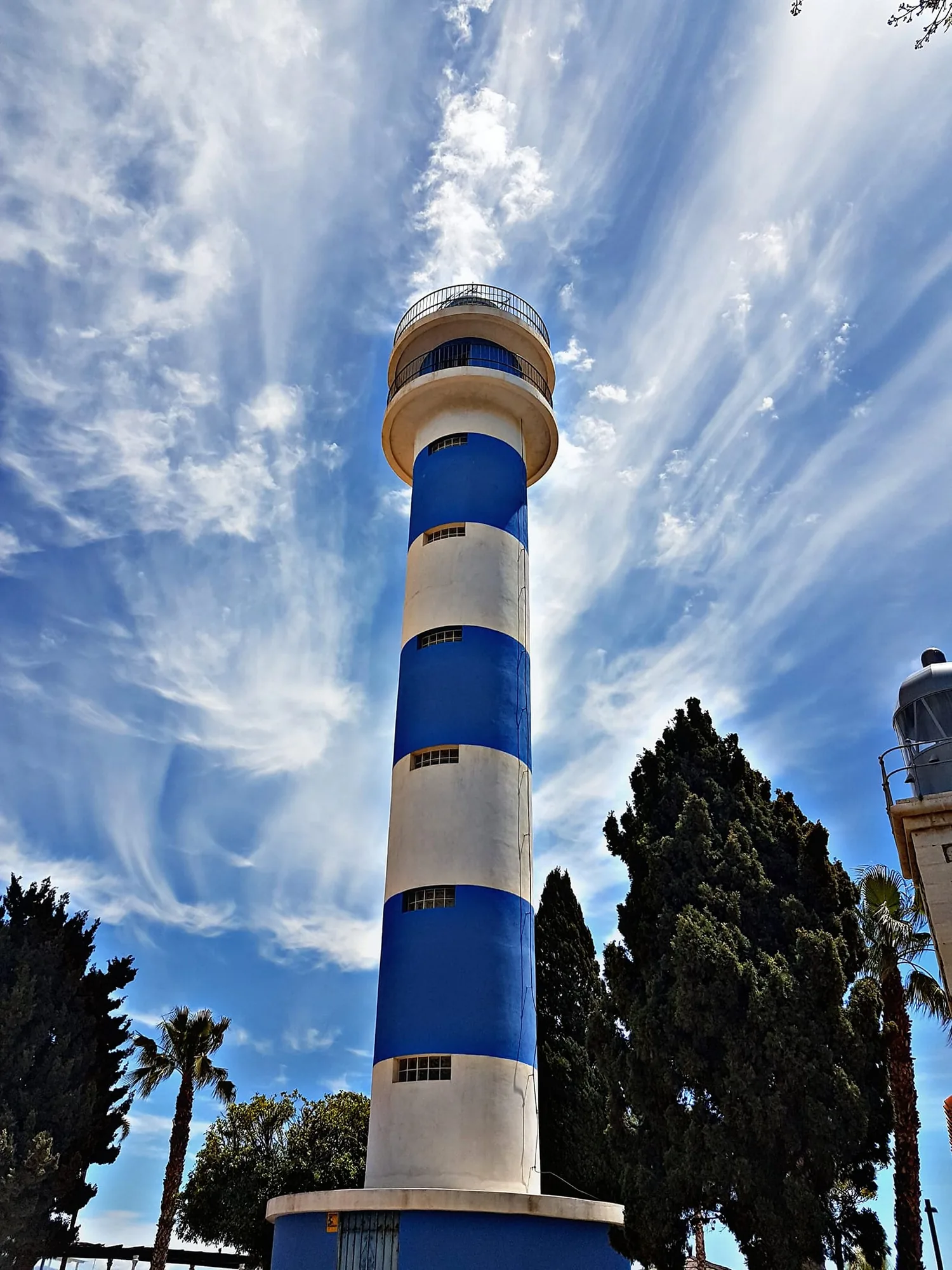 Faro de franjas azules y blancas con palmeras y un cielo de nubes finas en Torre del Mar