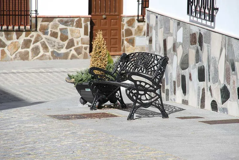 Banco de forja negro en una plaza con muros de piedra y una puerta de madera