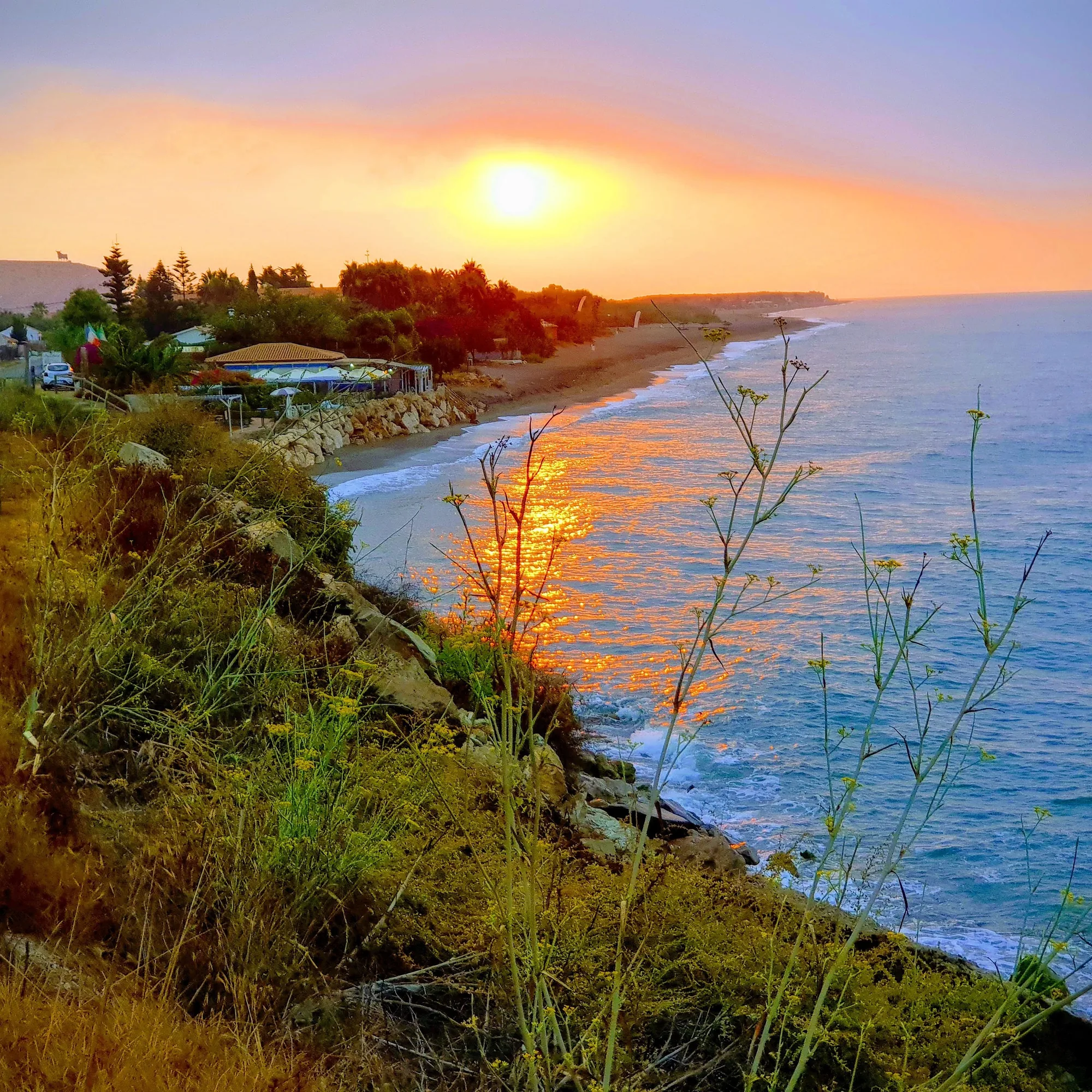 Sol sobre el mar al atardecer con la costa y una playa con chiringuito en Valle-Niza