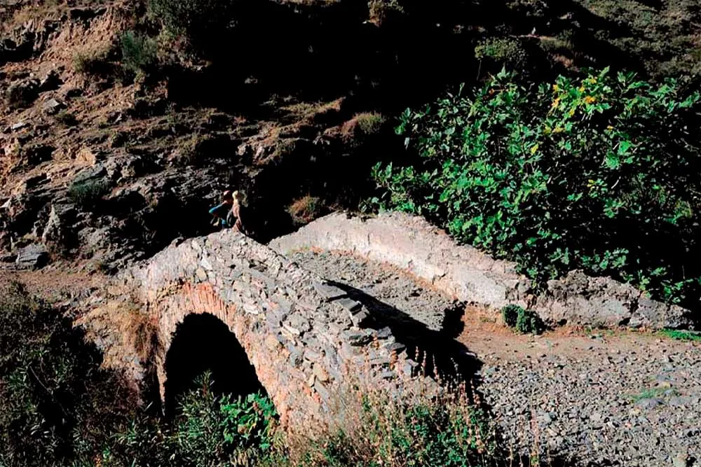 Pequeño puente de piedra de un solo arco sobre un sendero entre vegetación en Sedella