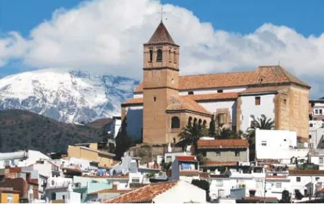 Iglesia de torre con tejado de teja sobre un pueblo blanco y montañas nevadas al fondo